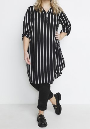 Persoon in een zwart-witte, verticaal gestreepte tuniek, zwarte opgerolde broek en zwarte loafers, met één voet over de andere gekruist staand.