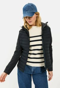 Zwarte gewatteerde jas met een capuchon, gecombineerd met een crèmekleurige gestreepte trui. Model draagt een denim pet en een blauwe spijkerbroek.