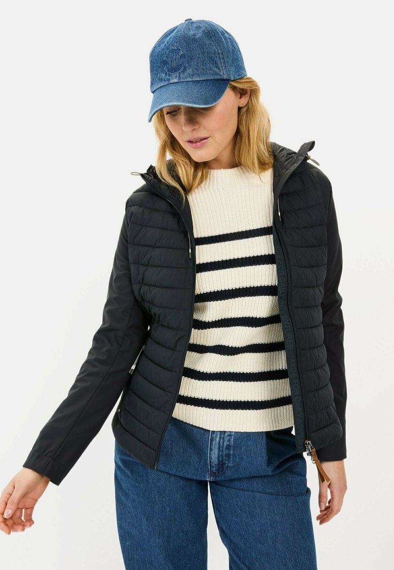 Zwarte gewatteerde jas met een capuchon, gecombineerd met een crèmekleurige gestreepte trui. Model draagt een denim pet en een blauwe spijkerbroek.