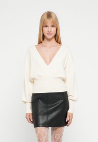 Pull-over crème à col en V avec manches bouffantes, associé à une mini-jupe en cuir noire et des collants blancs à motifs. Texture lisse dans l'ensemble.