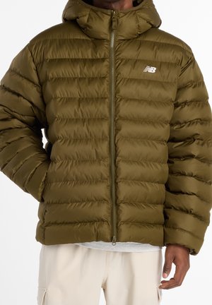 Homme portant une doudoune vert olive avec capuche, fermeture éclair sur le devant et logo New Balance sur la poitrine, associée à un pantalon beige clair.