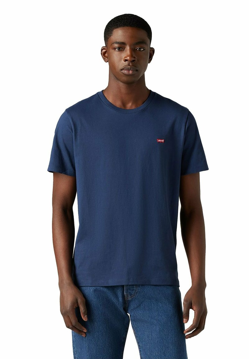 Młody mężczyzna ubrany w gładką, granatową koszulę Levi's i niebieskie dżinsy, stojący z rozluźnionymi rękami na białym tle.