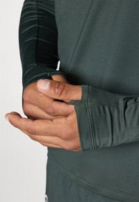 Cămașă sport cu mâneci lungi din țesătură verde închis, având găuri pentru degete la manșete și o textură netedă. Prim-plan al mâinilor pe țesătură.