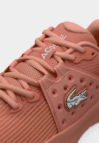 Κοντινό πλάνο σε αθλητικό παπούτσι κοραλί με υφασμάτινη υφή, κορδόνια και το λογότυπο κροκόδειλου της Lacoste στην πλευρική επιφάνεια.