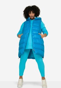 Modrá péřová vesta s kapucí, s prošívaným designem a bočními kapsami, nošena přes odpovídající modrý outfit s legínami a bílými teniskami.