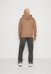 Felpa marrone con cappuccio e tasca a marsupio, vestibilità rilassata, polsini a coste, abbinata a un paio di jeans grigi scuri e larghi.