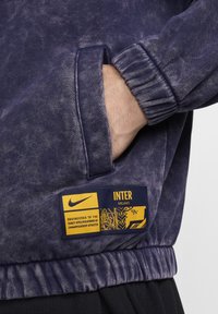 Nike-huppari purppurassa tie-dye-kankaassa, jossa on ribattu hihansuu, sivutasku ja keltainen etiketti, jossa lukee "INTER MILANO" sekä hoito-ohjeet.