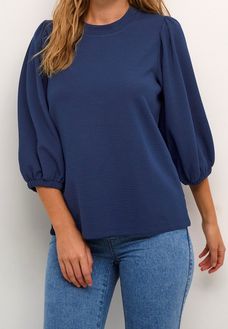 Top blu navy con finitura testurizzata, scollatura rotonda e maniche a sbuffo. Indossato con jeans blu chiaro, che evidenziano lo stile casual.