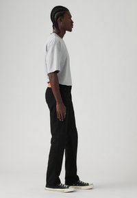 Muškarac s pletenom kosom nosi sivi oversized majicu, crne ravne hlače i crne niske tenisice i stoji bočno na jednobojnoj pozadini.