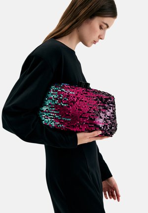 Donna con un vestito nero che tiene una pochette rettangolare coperta di paillettes lucide rosa e turchesi su uno sfondo semplice.