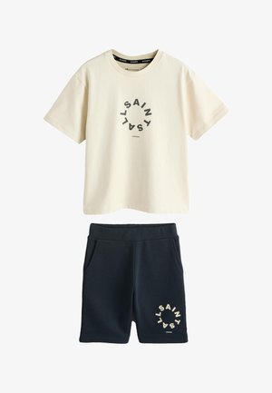 Jongen staat, draagt een beige oversized T-shirt en zwarte korte broek met cirkelvormig tekstontwerp, witte sneakers en sokken, tegen een effen lichte achtergrond.