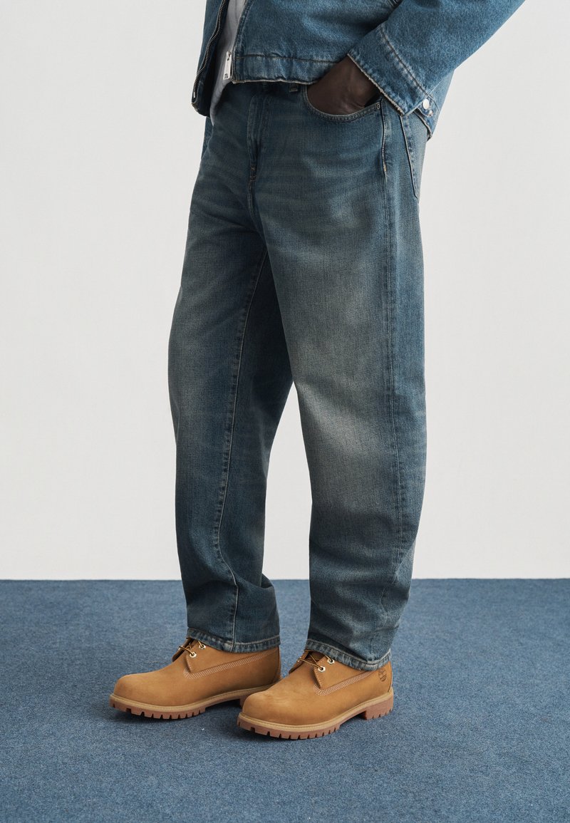 Personne portant un jean bleu délavé, des bottes en cuir marron clair à lacets, et une veste en jean bleue, une main dans la poche, debout sur un tapis bleu.