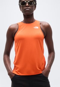 Άτομο που φορά ανακλαστικά γυαλιά ηλίου και πορτοκαλί αμάνικο αθλητικό τοπ The North Face, με τα χέρια στις τσέπες ενός μαύρου σορτς.