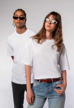 Two models wear loose-fitting white t-shirts. One has black sunglasses, the other features silver-accented sunglasses. The woman wears blue jeans with a brown belt.