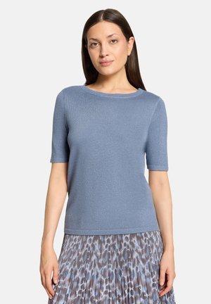 Femme aux cheveux bruns et raides portant un haut bleu clair à manches courtes et une jupe plissée avec un imprimé animalier bleu et gris.