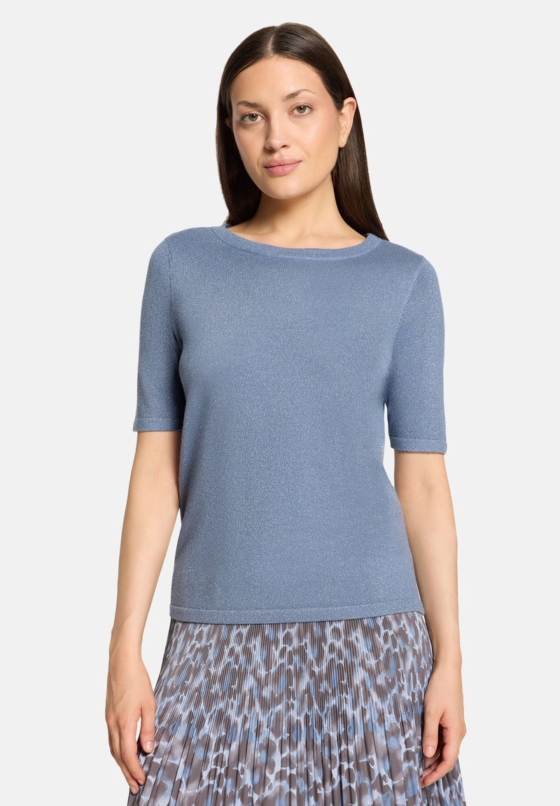 Femme aux cheveux bruns et raides portant un haut bleu clair à manches courtes et une jupe plissée avec un imprimé animalier bleu et gris.