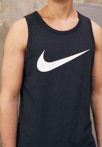 Svart ärmlös linne i bomull med en stor vit Nike swoosh-logotyp på bröstet. Slät textur med rund halsringning.