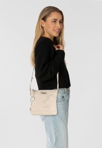 Beige Umhängetasche mit geprägtem Design, silbernem Beschlag und abnehmbarem Anhänger. Glatte Textur und verstellbarer Riemen.