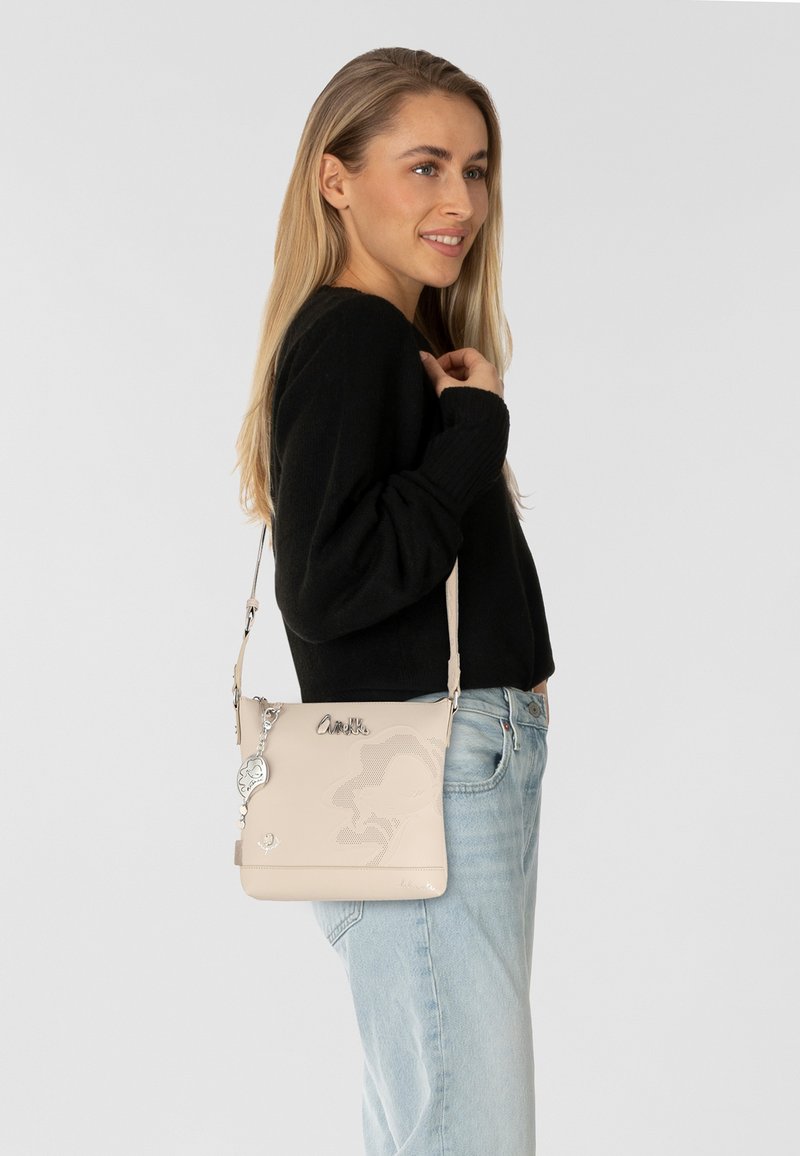 Beige Umhängetasche mit geprägtem Design, silbernem Beschlag und abnehmbarem Anhänger. Glatte Textur und verstellbarer Riemen.