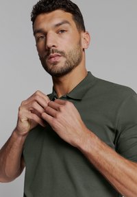 Donkergroene polo met een textuurstof, voorzien van een knoopsluiting en een klassieke kraag. Handen die de kraag aanpassen zijn zichtbaar.