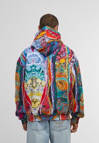 Mann trägt einen bunten, gemusterten Kapuzenpullover mit abstrakten, geometrischen Designs und hellblauen Jeans mit gesticktem Logo auf der Tasche, mit dem Rücken zur Kamera.