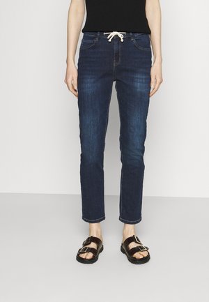 Persoon draagt donkerblauwe denim jeans met een witte trekkoord en zwarte sandalen met twee gespen, staand tegen een eenvoudige lichte achtergrond.