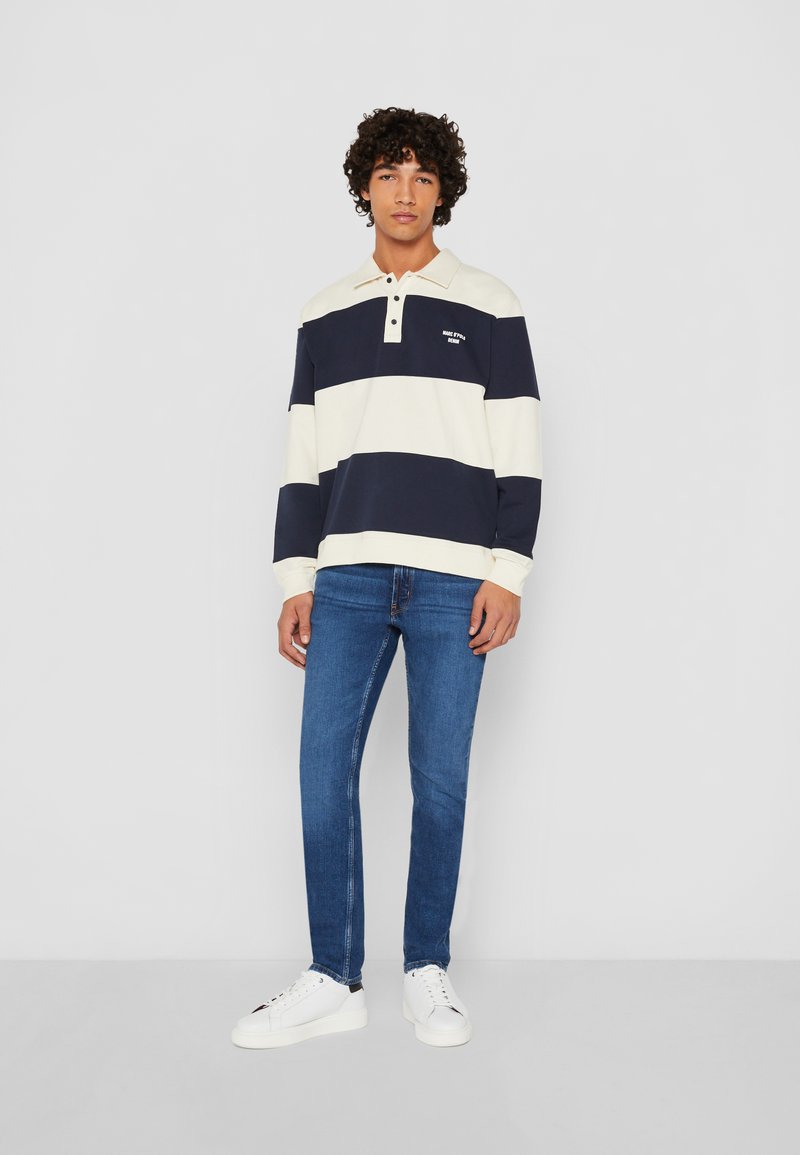 Polo à manches longues rayé bleu marine et crème avec un col, trois boutons et un petit logo. Porté avec un jean slim-fit bleu et des baskets blanches.