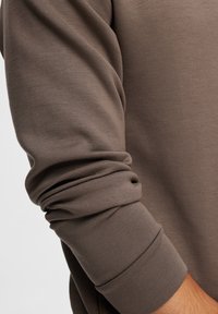 Langsleeve top in donker taupe stof, met een gladde textuur en aansluitende manchetten. Het materiaal lijkt zacht en rekbaar.