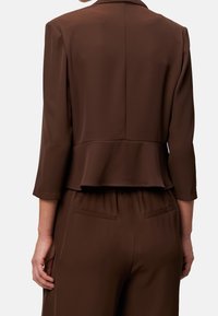 Ruskea lyhyt bleiseri, jossa on kolme neljänneshihaa, sileä pinta ja volangiyksityiskohta, yhdistetty samansävyisiin ruskeisiin housuihin sivutaskuilla.