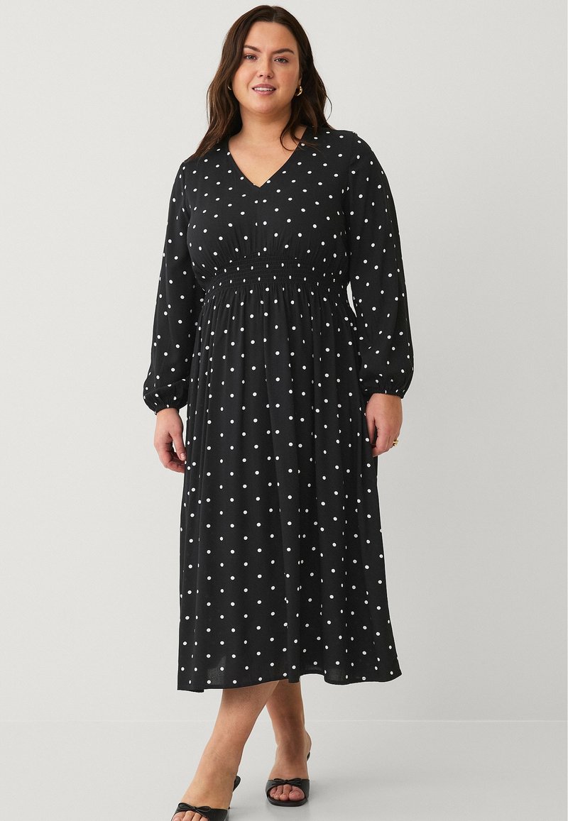 Schwarzes, mittelange Kleid mit weißen Punkten, V-Ausschnitt, langen Ärmeln, gerafftem Bund und fließendem Rock, aus leichtem Stoff gefertigt.