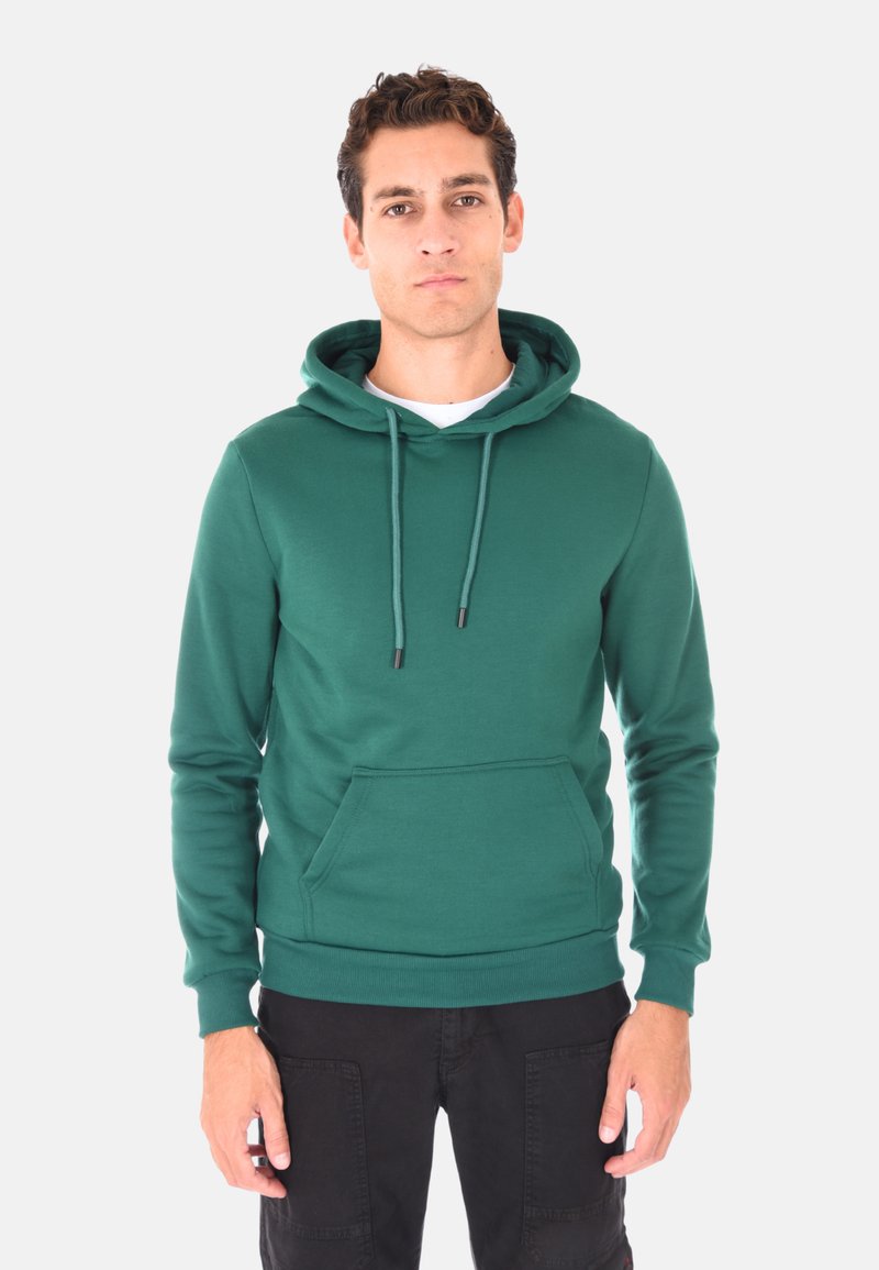 Uomo in piedi che indossa un felpa con cappuccio verde con tasca frontale e pantaloni neri, su uno sfondo bianco semplice.