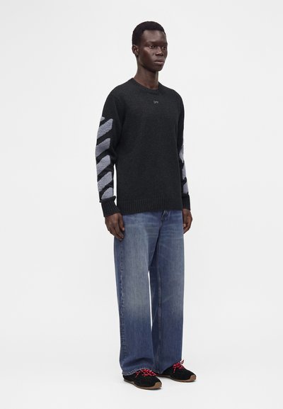 Homme portant un pull gris foncé avec des rayures diagonales sur les manches, un jean bleu large et des chaussures noires avec des lacets rouges, debout devant un fond blanc.