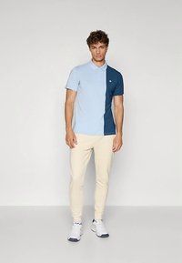 Man in een lichtblauwe en donkerblauwe polo, beige broek en witte sneakers, staand tegen een neutrale achtergrond.