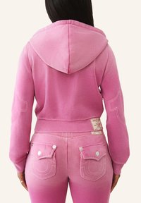 Hoodie rose avec capuche et détails de couture, associé à un pantalon de jogging rose dégradé avec poches arrière et accents de boutons.
