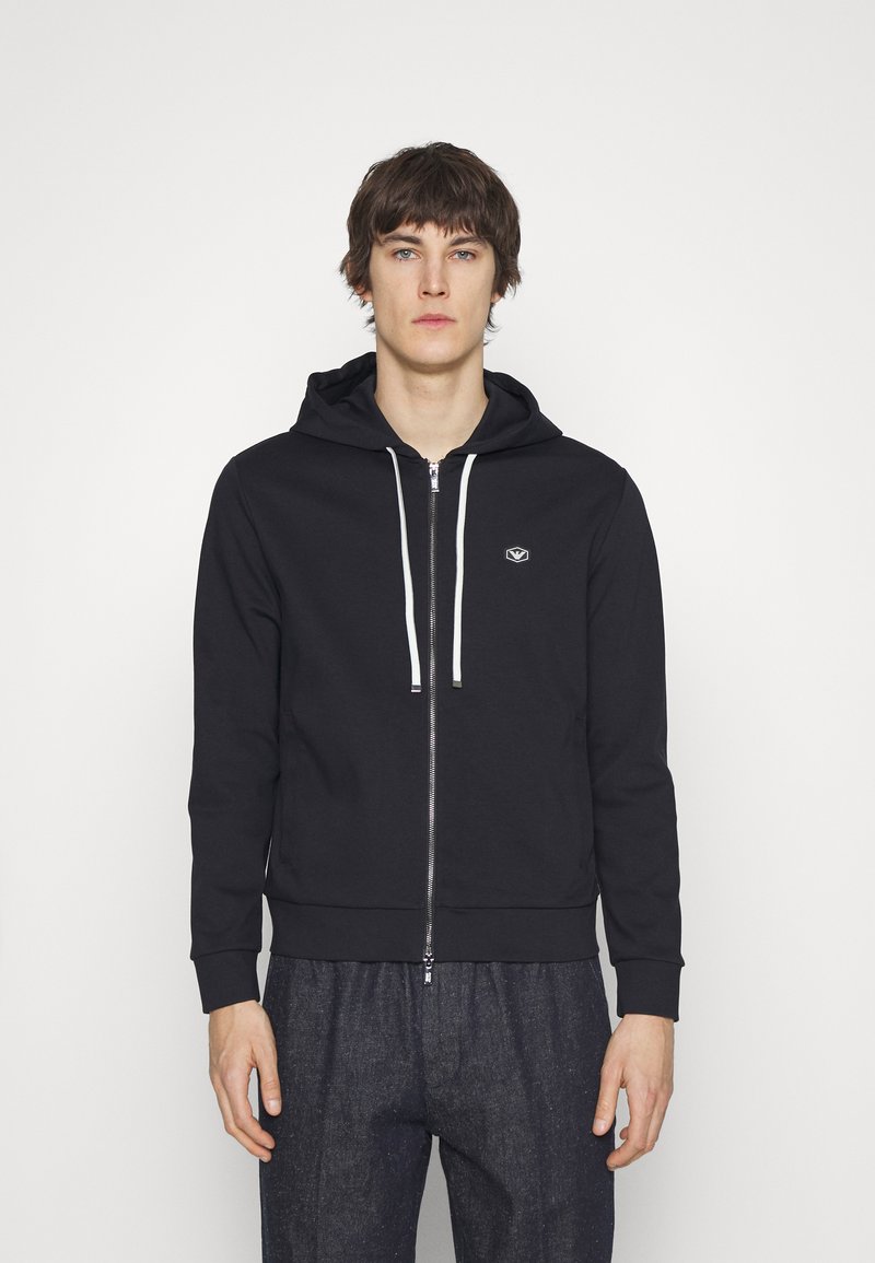 Black zip-up hoodie with a drawstring hood, featuring a small logo on the chest, paired with loose, dark patterned pants.