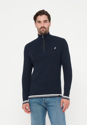 Homme portant un pull côtelé bleu marine à demi-fermeture éclair avec des poignets et un ourlet rayés blancs, associé à un jean bleu clair sur fond blanc.