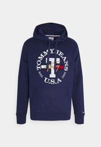 Námořnická modrá mikina z bavlny, se světlým velkým potiskem loga a barevnými akcenty. Obsahuje klokaní kapsu a kapuci se stahovací šňůrkou.
