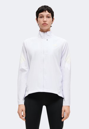 Personne aux cheveux courts et foncés portant une veste blanche zippée et un pantalon noir, debout devant un fond clair uni.