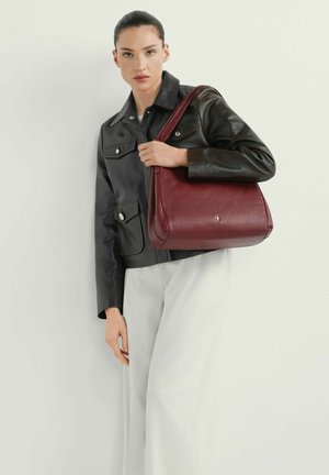 Kobieta w czarnej skórzanej kurtce i białych spodniach, trzymająca dużą bordową skórzaną torbę na ramię na jednolitym tle.