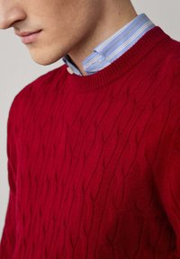 Pull en maille rouge, col rond, avec un motif en relief. En dessous, une chemise à col rayée bleu clair est visible.