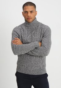 Jersey de cuello alto gris de punto con un patrón de trenzas texturizadas, puños y dobladillo acanalados. Llevado con pantalones azul marino. Silueta ajustada.