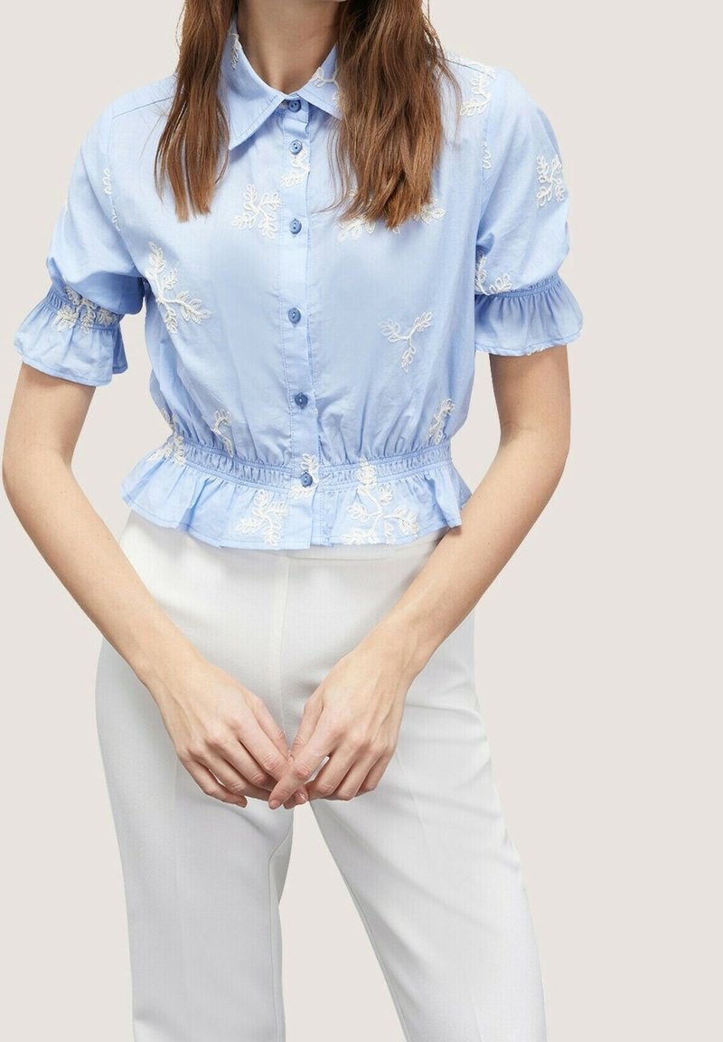 Light blue short-sleeve blouse with embroidered floral patterns, button closure, and elasticized waist. Worn with white trousers.