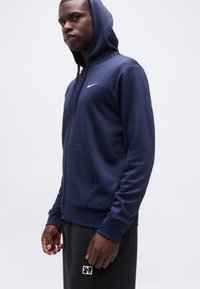 Νέος άνδρας με μπλε σκούρο φούτερ Nike με φερμουάρ και κουκούλα καλυμμένη, και σκούρο γκρι παντελόνι φόρμας Nike, σε απλό ανοιχτό φόντο.