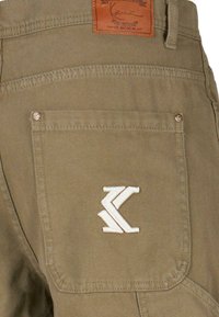 Pantalons vert olive avec une poche arrière arborant un logo blanc brodé. Comprend une étiquette en cuir marron au-dessus de la poche. Texture de tissu durable.