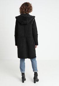 Abrigo negro con capucha, hecho de un material texturizado y suave, con costuras visibles y un corte largo, combinado con unos vaqueros arrollados de color azul claro y botas negras.