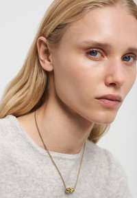 Guldkæde med vedhæng i form af en kugle, båret over en lysegrå, tekstureret sweater. Nærbillede af en persons ansigtstræk.