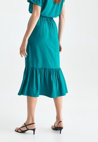 Robe teal jusqu'au genou avec une taille froncée et un ourlet évasé, associée à des talons noirs à brides. Tissu lisse avec une texture minimale.