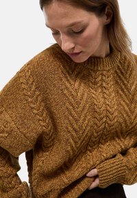Pull en tricot de couleur jaune moutarde chaud, présentant un motif en cable texturé, un col et des poignets côtelés, avec une coupe ample et une texture douce.