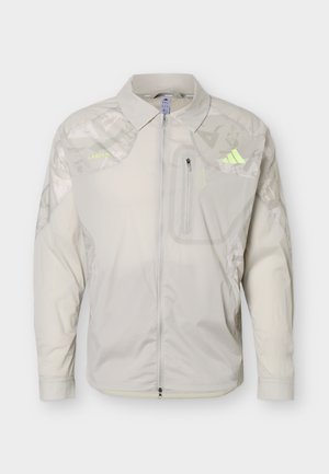 Chaqueta beige claro con cremallera, cuello, bolsillo con cremallera en el pecho, sutil estampado de camuflaje en los hombros y mangas, y detalles de marca en amarillo neón.
