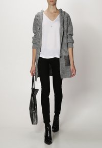 Cardigan grigio lavorato a maglia con cappuccio, top bianco a scollo a V, pantaloni neri skinny e stivaletti neri lucidi, mentre tiene una borsa tote nera.